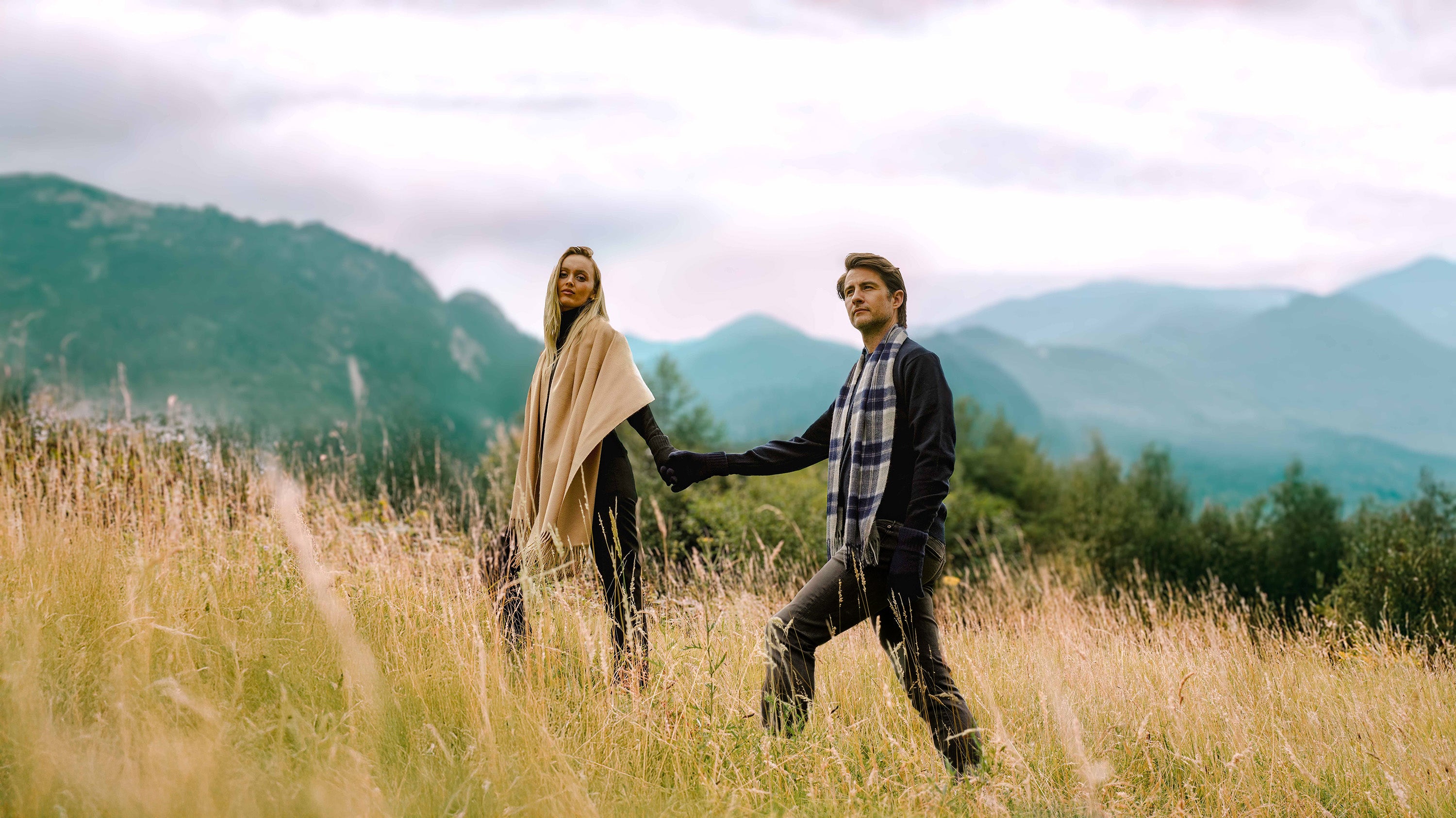 Two Models walking hand-in-hand through a field with mountains in the background. Male model wearing Lona Scott Cashmere Scarf and Jumper and Female Model wearing Cashmere Shawl